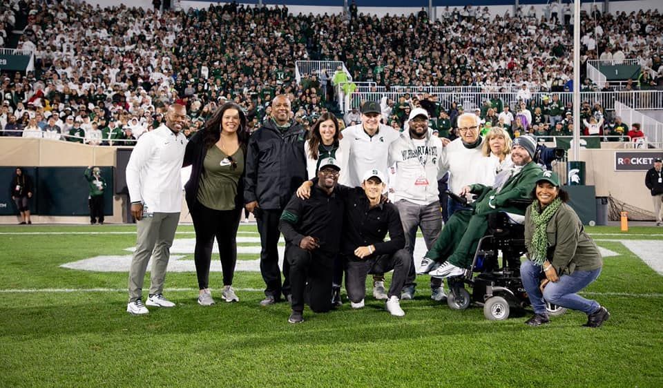 Group of people posing on a football field. Some wearing green, many smiling. Stadium crowd in the background.