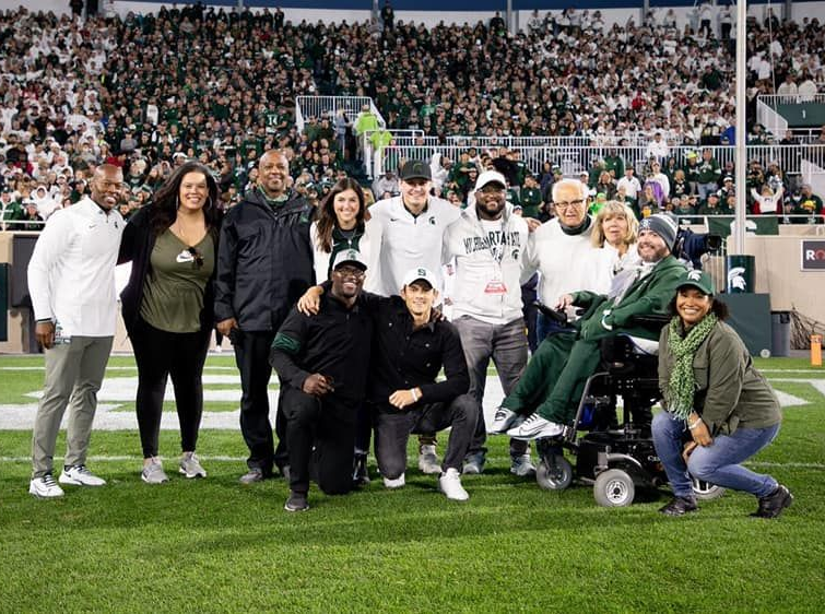 Group of people posing on a football field. Some wearing green, many smiling. Stadium crowd in the background.