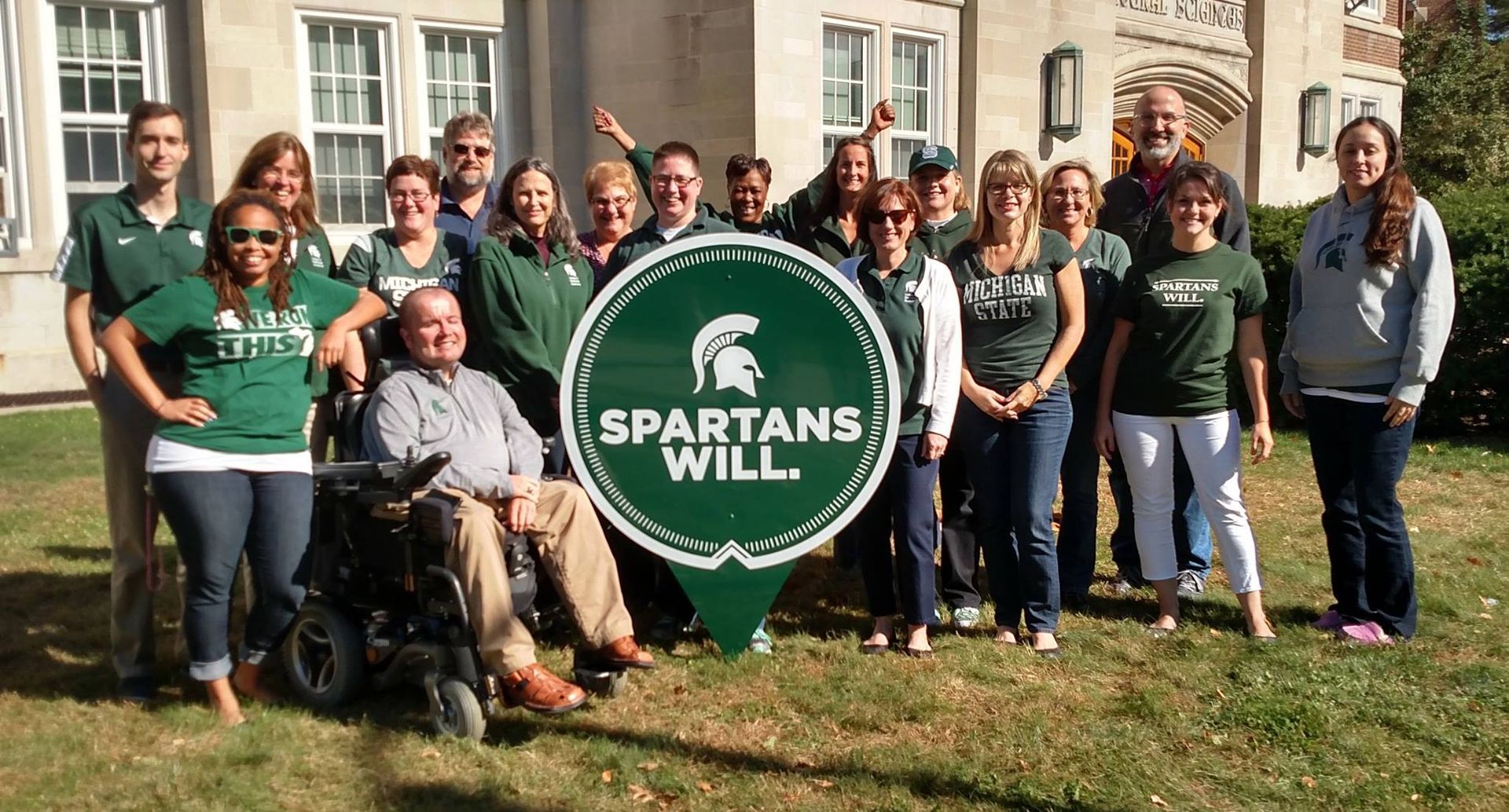 Group of people, many wearing green Spartan shirts, posing in front of a building with a “Spartans Will” sign.