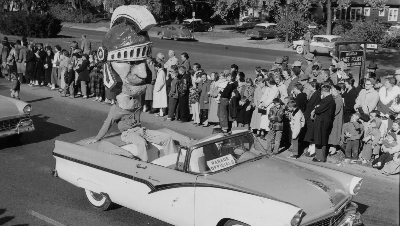 Parade float with giant knight head in convertible car, crowds line street.
