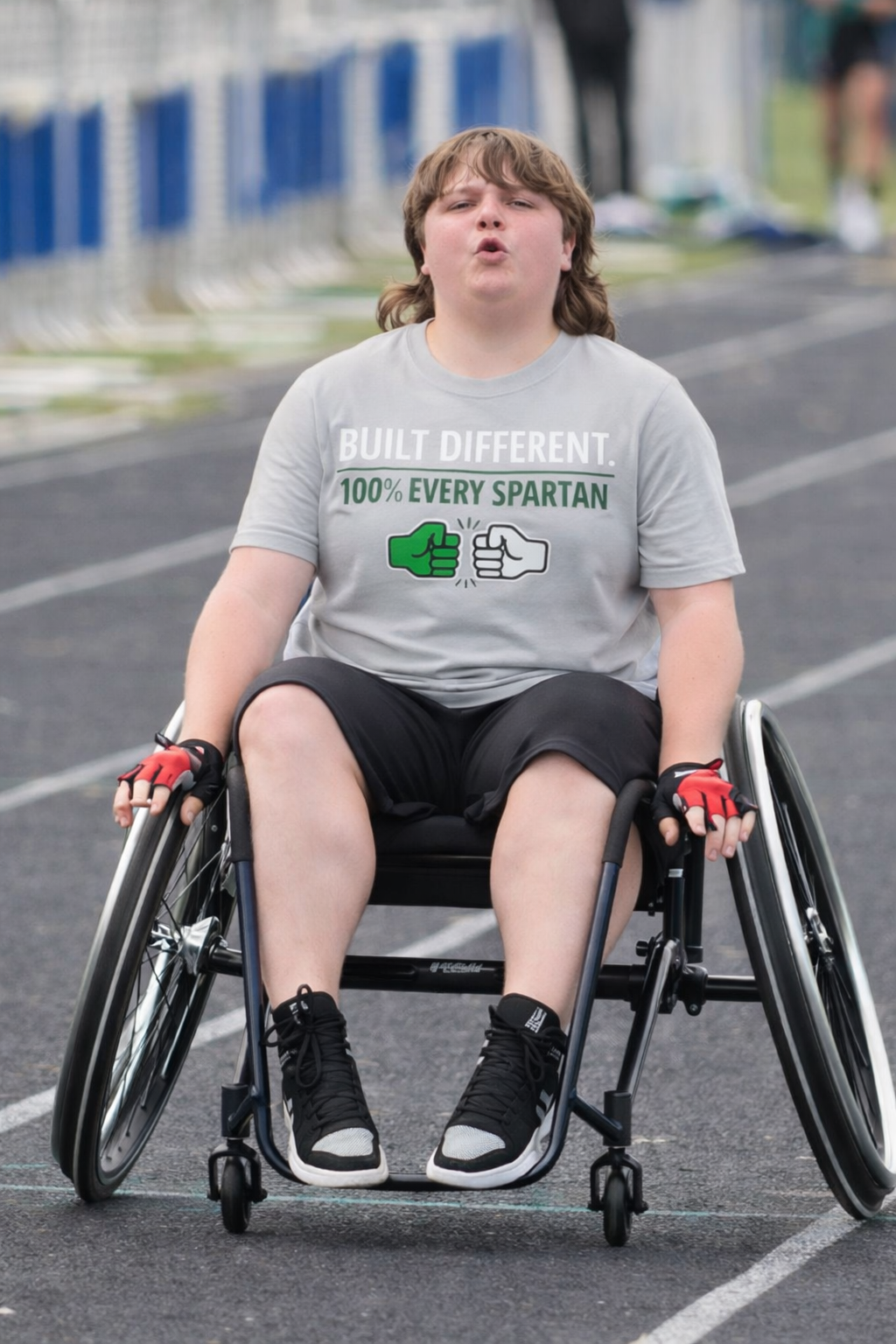 Person in a wheelchair on a track, wearing a gray adaptive Every Spartan Built Different short sleeve shirt and black shorts.