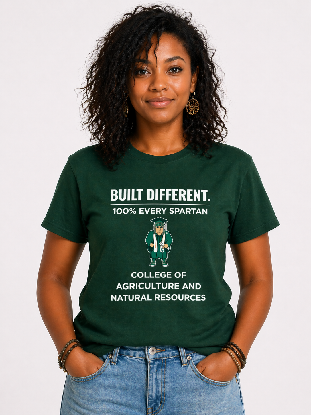 Person in a green “BUILT DIFFERENT” Spartan agriculture T-shirt and jeans, standing with hands in pockets