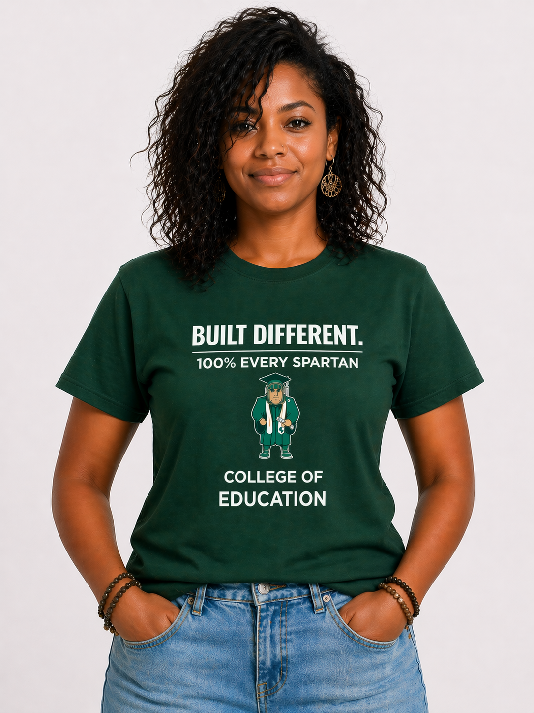 Person wearing a green “BUILT DIFFERENT” College of Education T-shirt and jeans, standing against a white background.