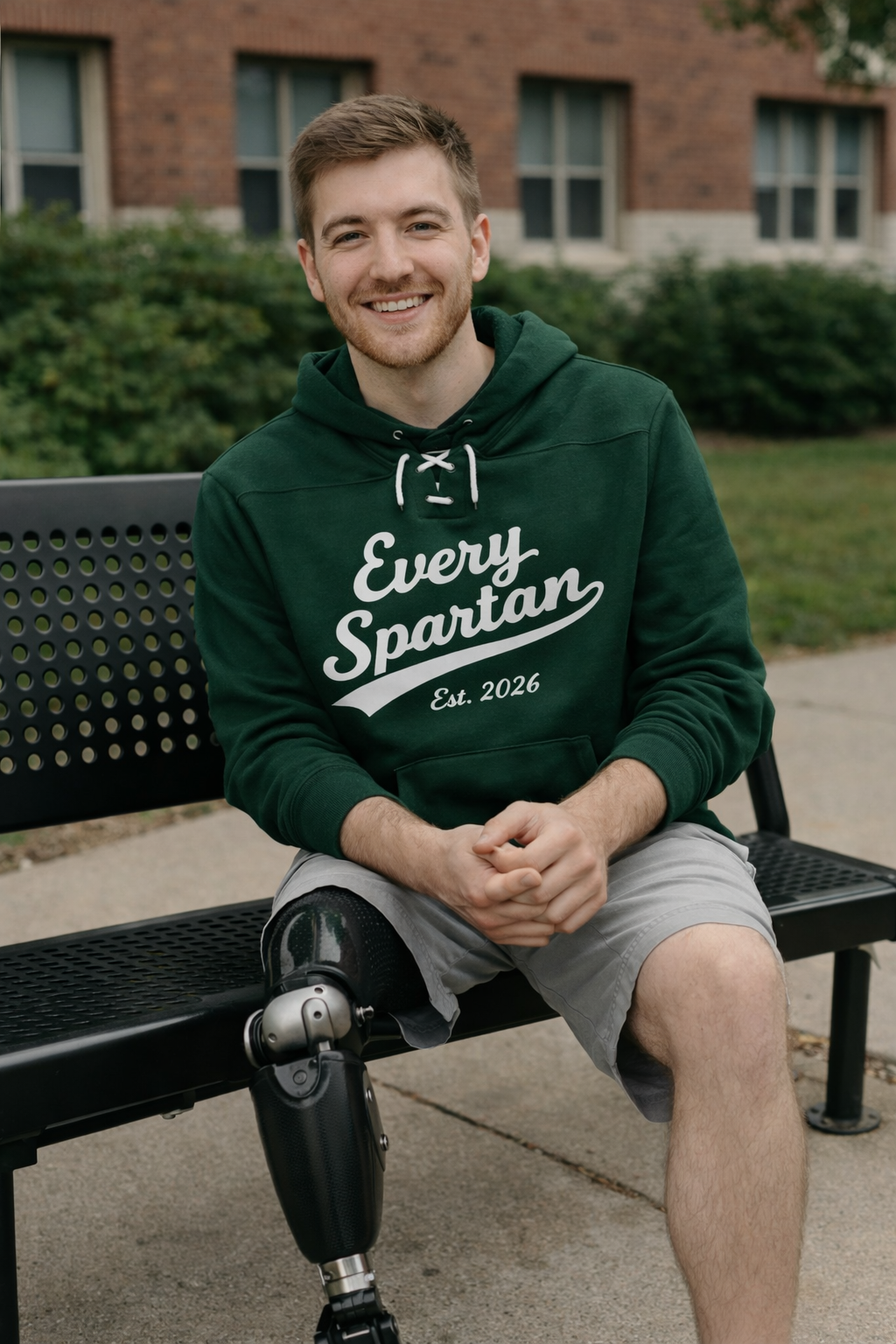 Man with prosthetic leg wearing green hoodie that says “Every Spartan,” sitting on a park bench.