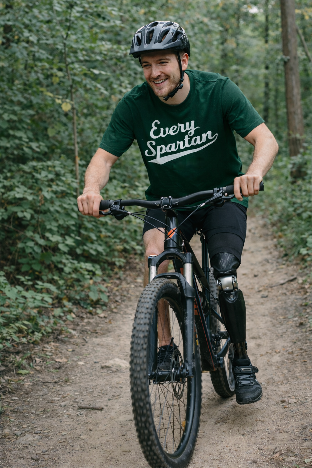 Man with prosthetic leg rides mountain bike on a dirt path, smiling. Forest background, wearing helmet and green shirt.