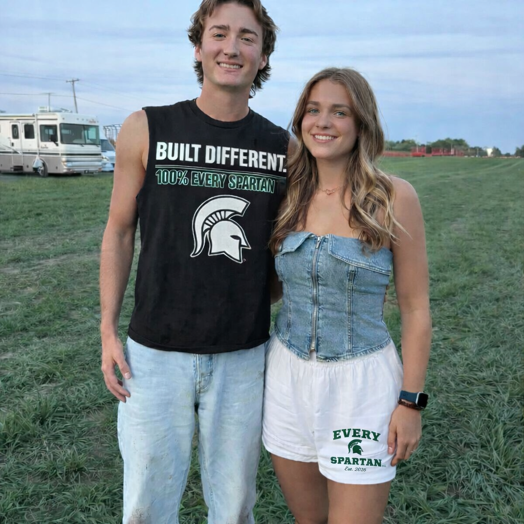 Two people standing outdoors, smiling. Man in a tank top, woman in a denim top and shorts. Green field, RV in background.