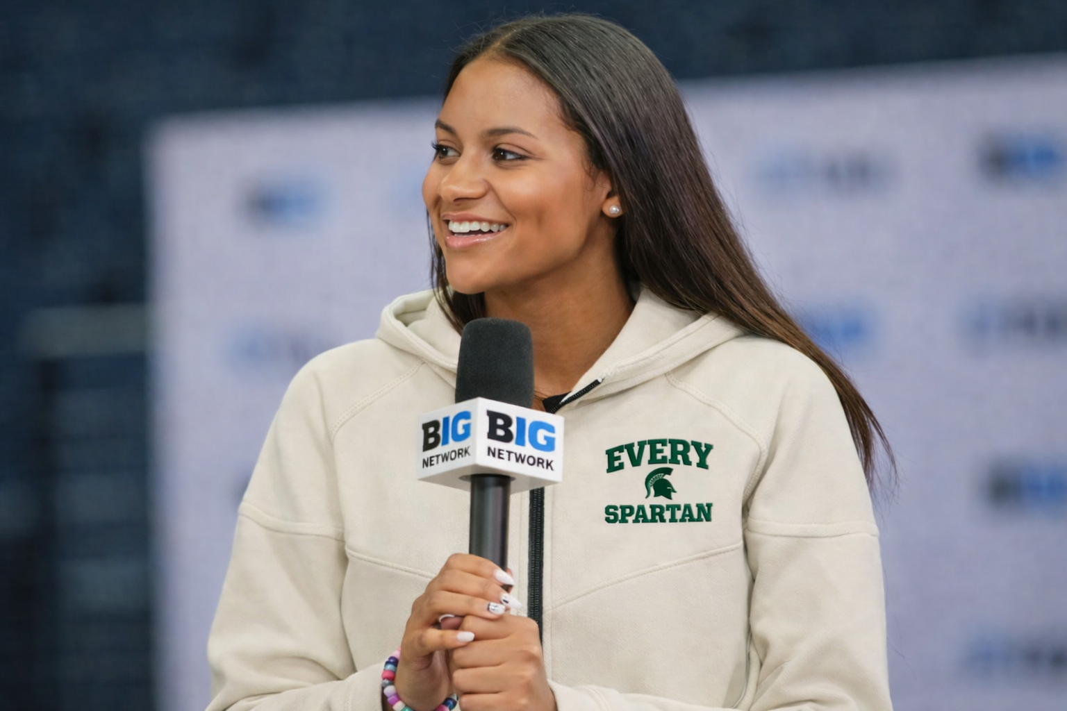 Woman holding a microphone, smiling. Wearing a cream-colored sweatshirt with 