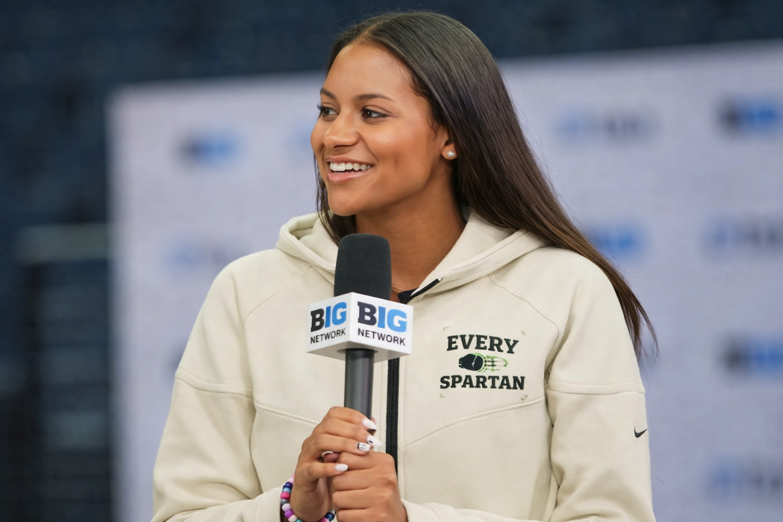 Woman holding a microphone, smiling. Wearing a hoodie with 