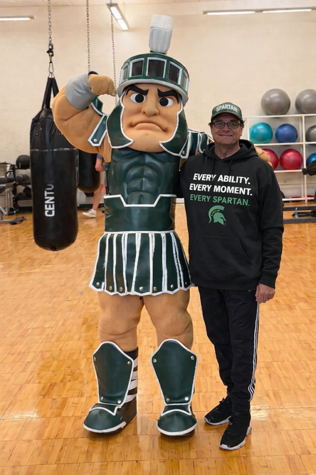 Spartan mascot flexes arm next to a person in Spartan apparel in a gym.