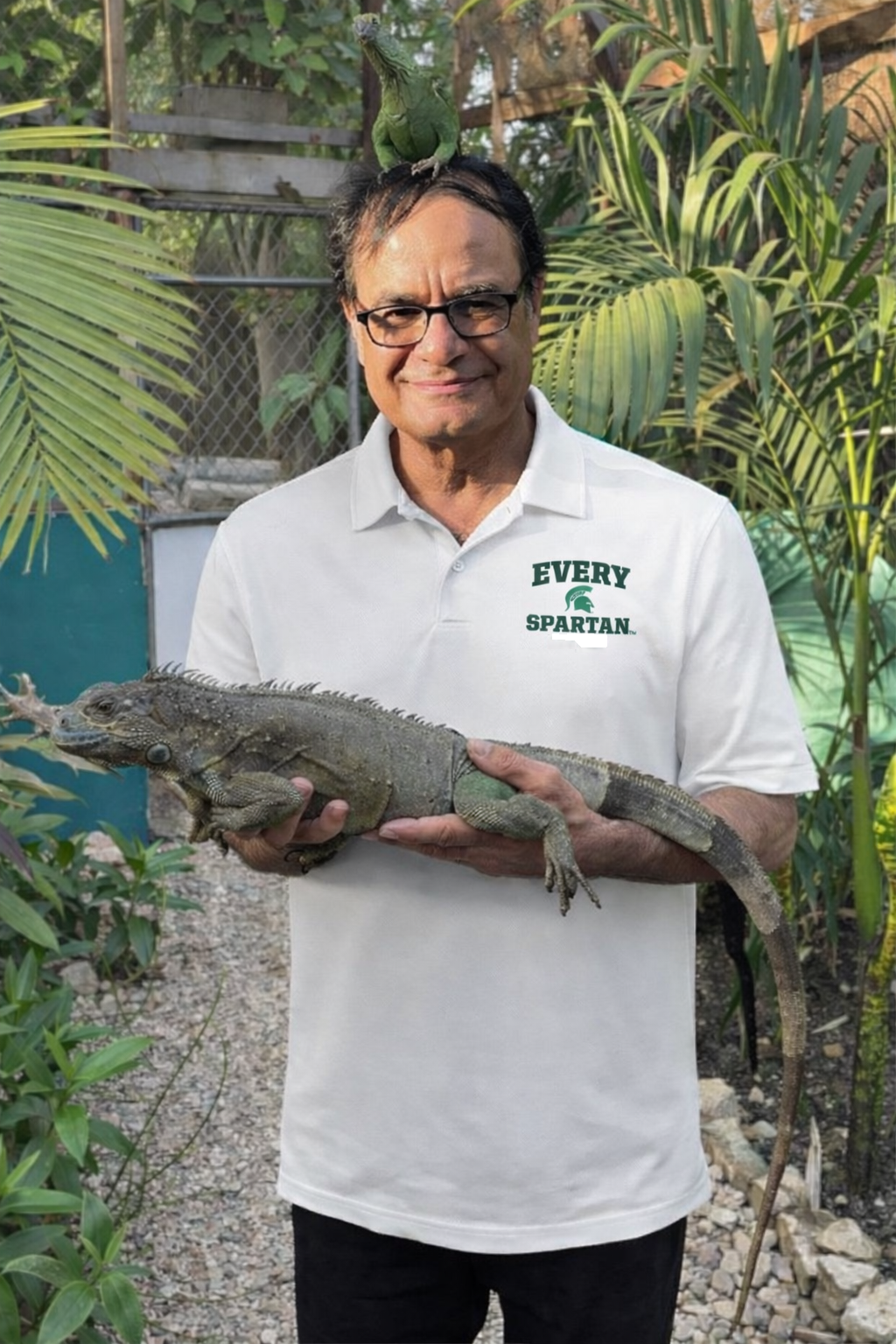 Man holding a large iguana outside; wearing a white polo shirt with 