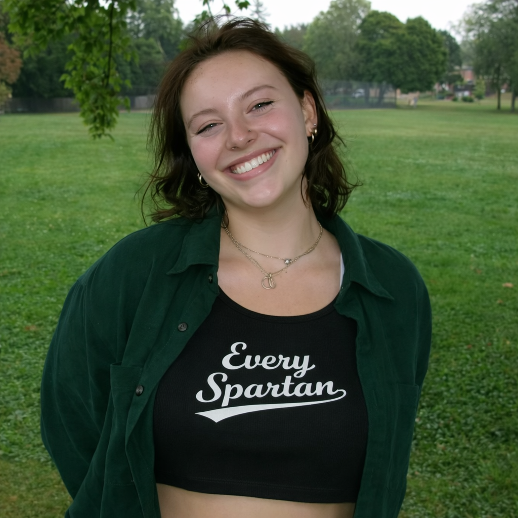 A smiling person outdoors, wearing a dark green shirt over a black crop top that says 