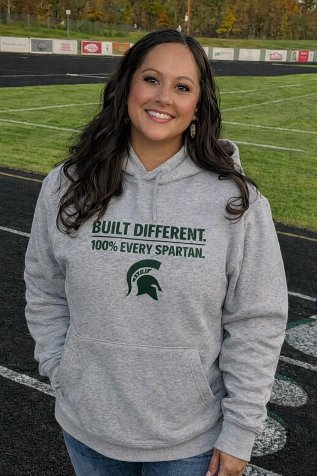 A smiling person stands on a sports field wearing a gray hoodie with 
