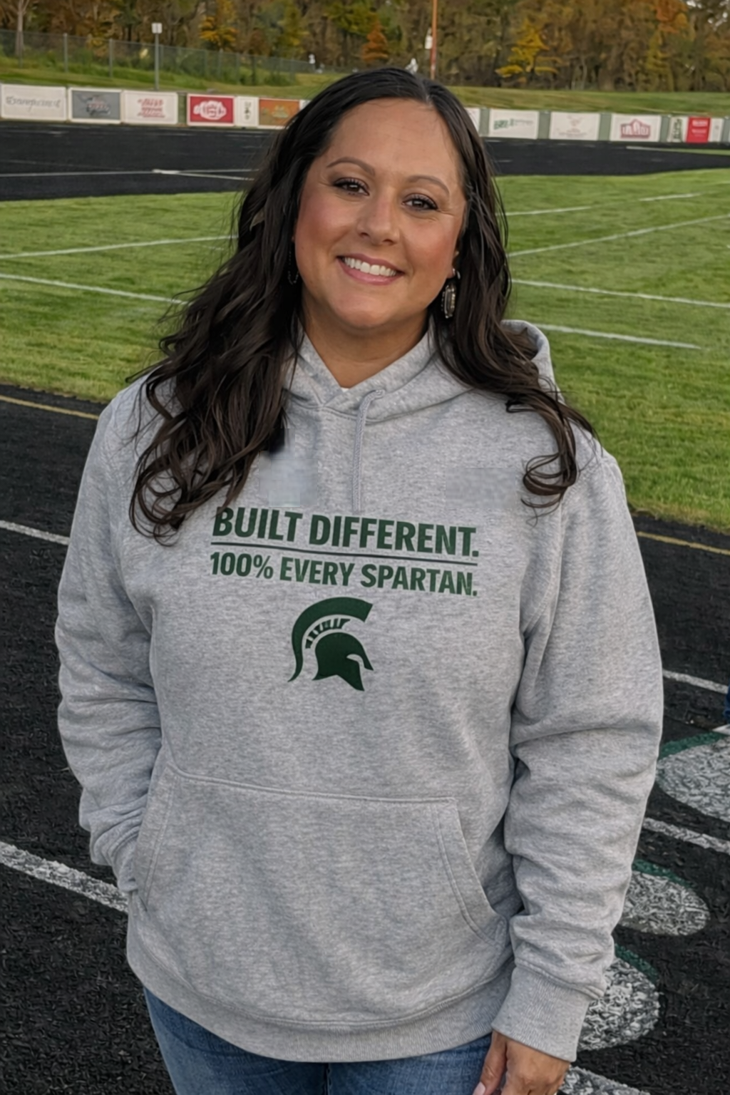 A smiling person wearing a gray hoodie with a green Spartan logo and text standing on a football field.