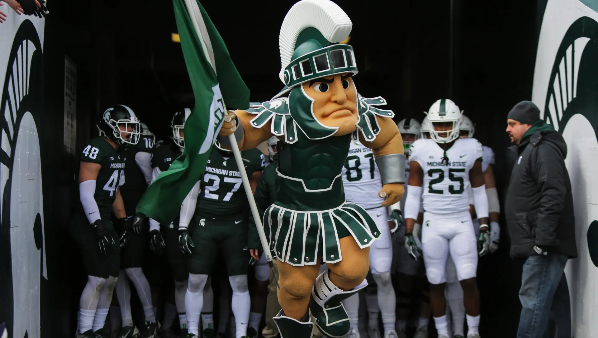 Spartan mascot leads football team from tunnel, green and white uniforms.