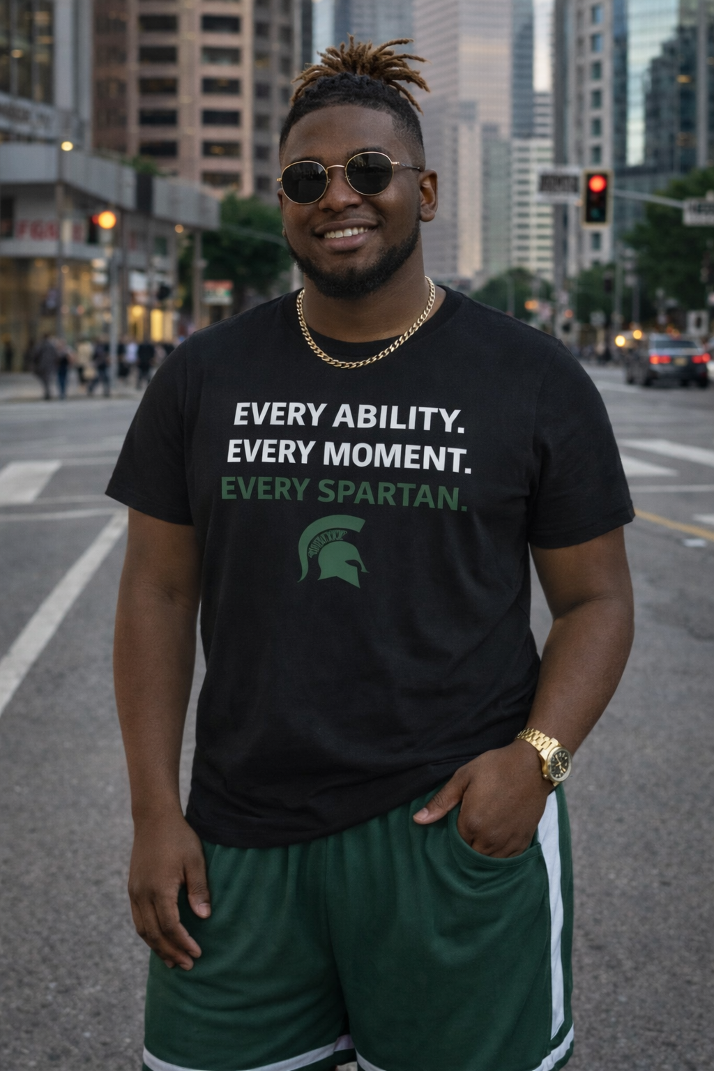 Man in black shirt and green shorts with Spartan logo, smiling outdoors on a city street.