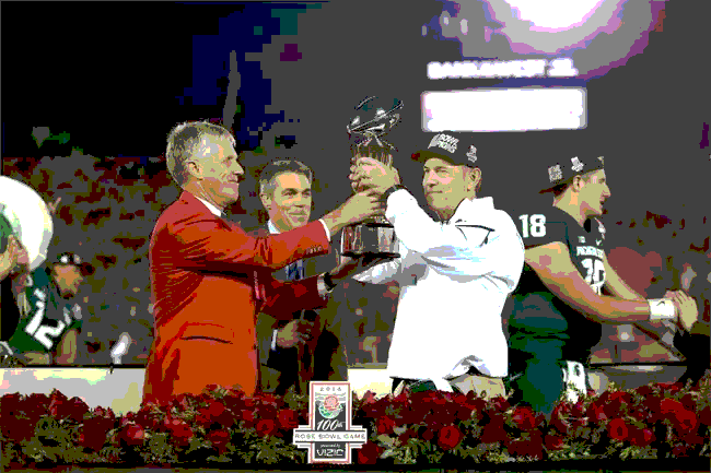 Men in formal wear and athletic gear hold a trophy on a platform with roses; football stadium in background.