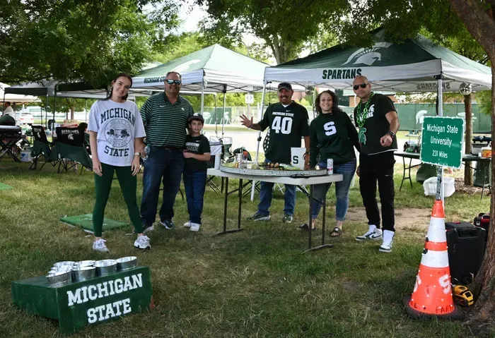 Group of people at a Michigan State tailgate. They are standing near tables, tents, and decorations, all in green and white.