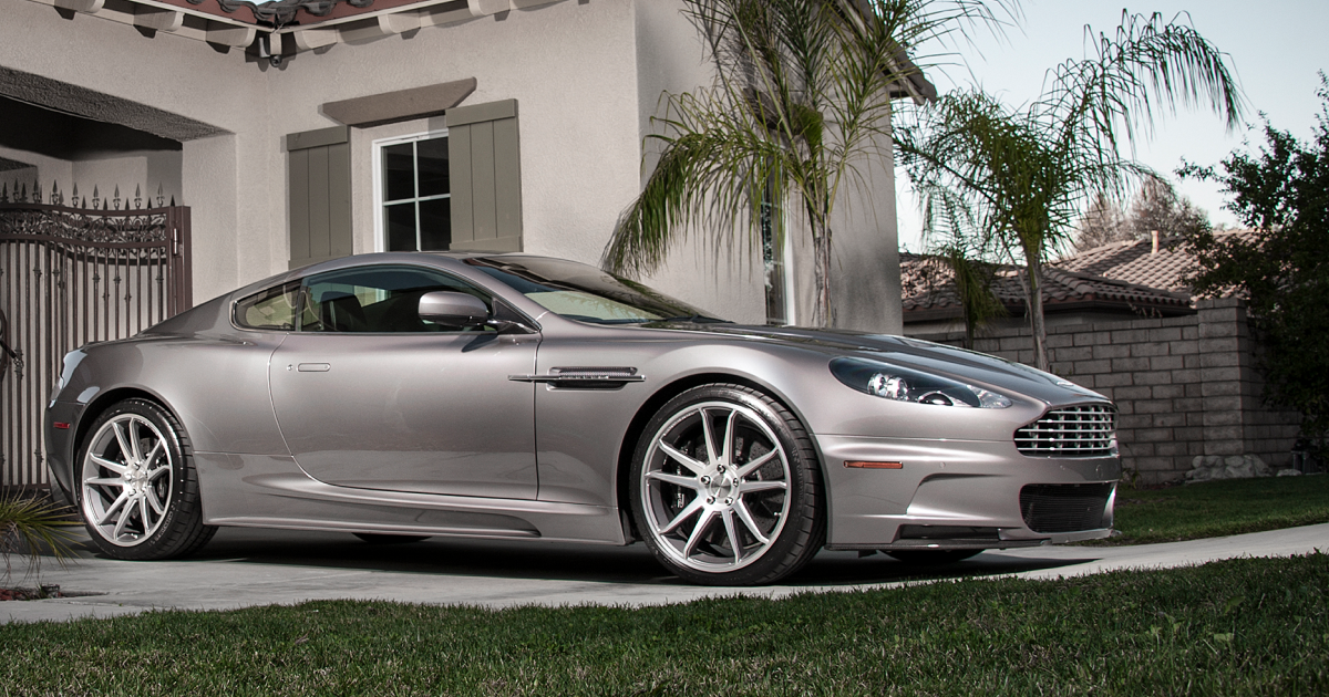 Silver Aston Martin coupe parked on a grassy driveway in front of a house.