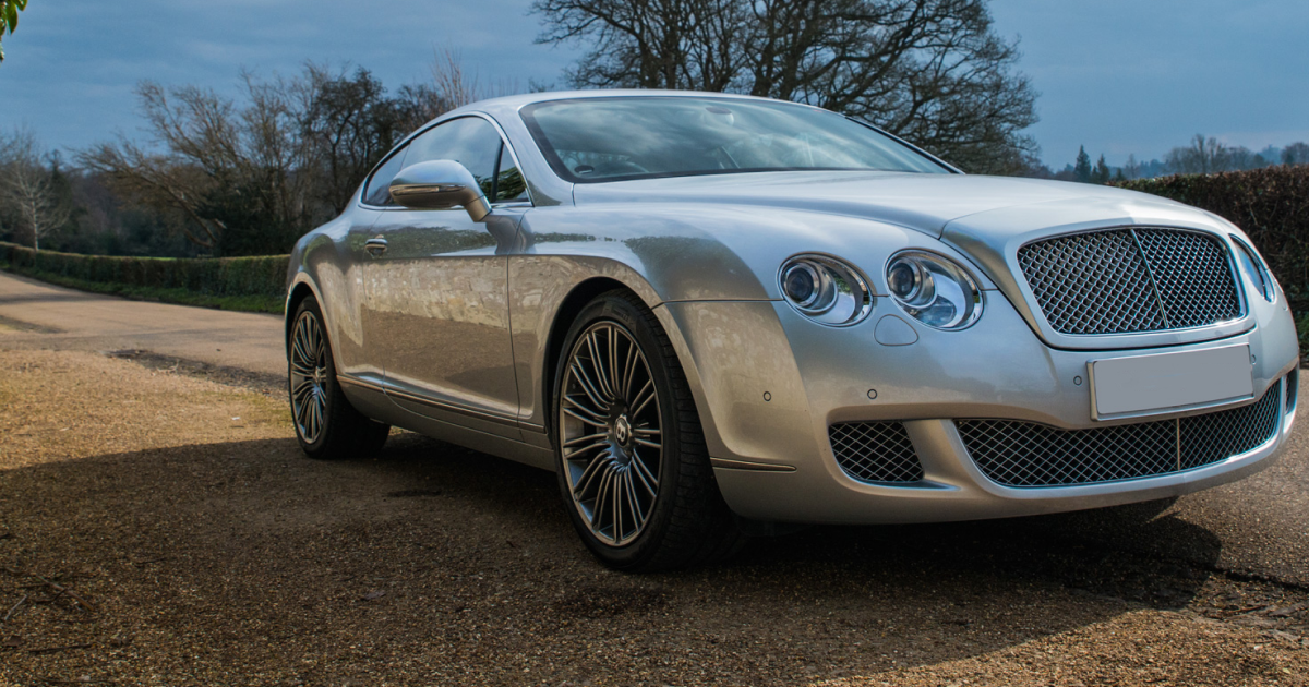 Silver Bentley coupe parked on gravel driveway, near a roadside hedge.