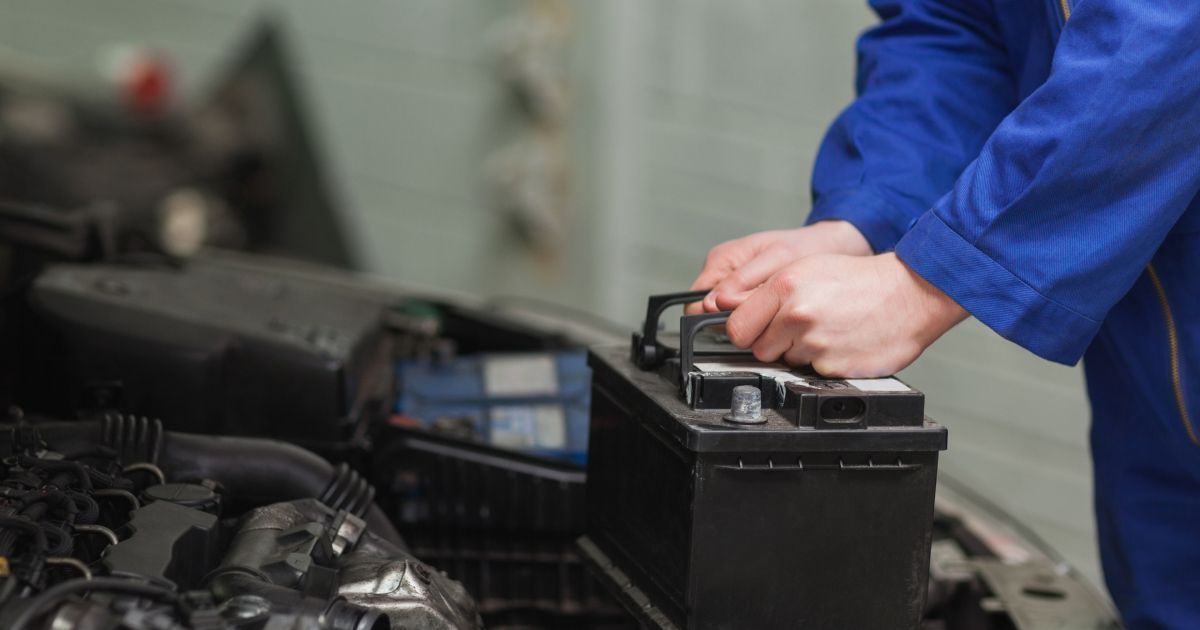 Person in blue overalls handling a car battery inside a vehicle's engine compartment.