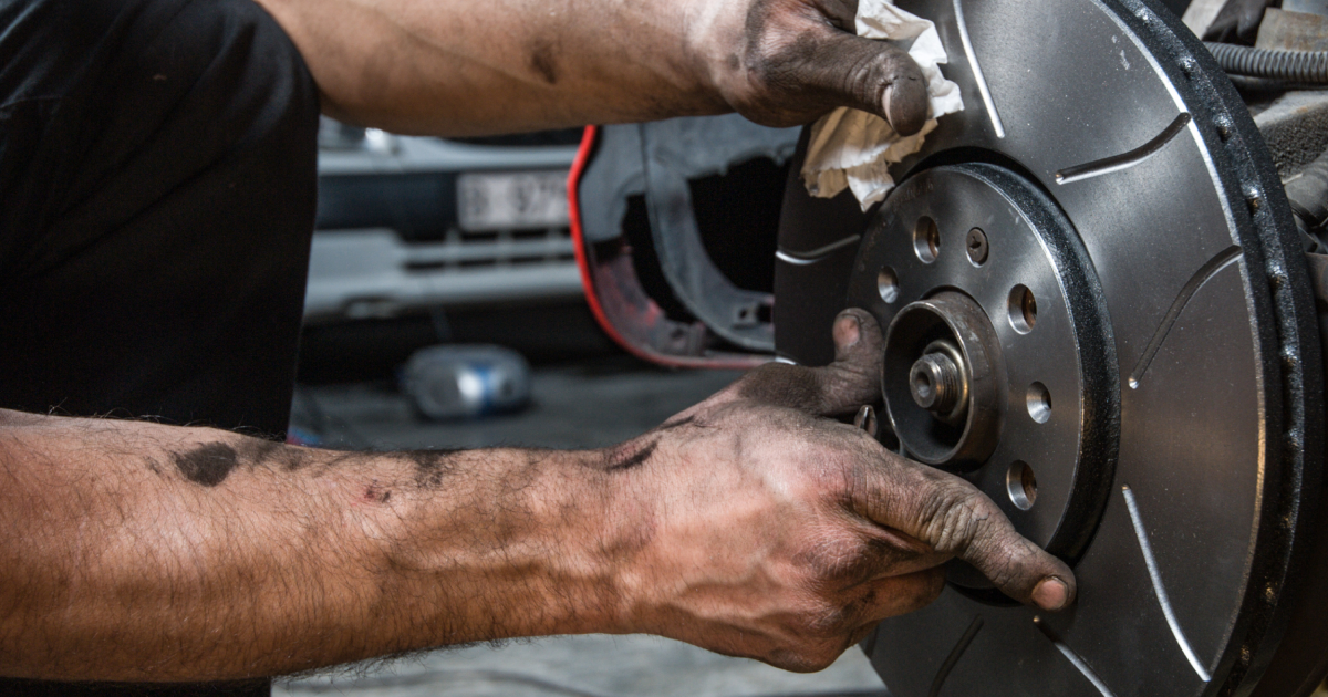 Mechanic's dirty hands working on a car's brake rotor in a garage setting.