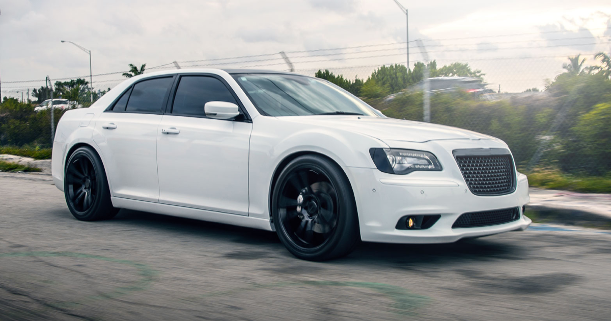 White Chrysler 300 sedan with black rims parked on a road. Cloudy day with trees in the background.