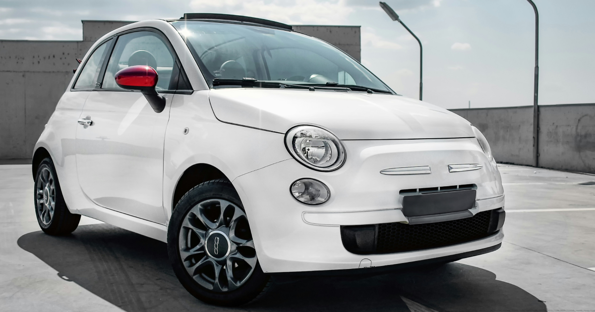 White Fiat 500 car with red side mirror parked on a rooftop parking lot with tall lampposts.