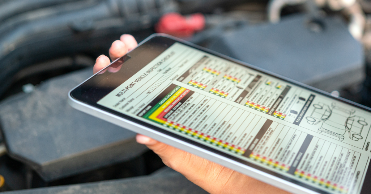 Hand holding a tablet with a car diagnostic report displayed near a car engine.