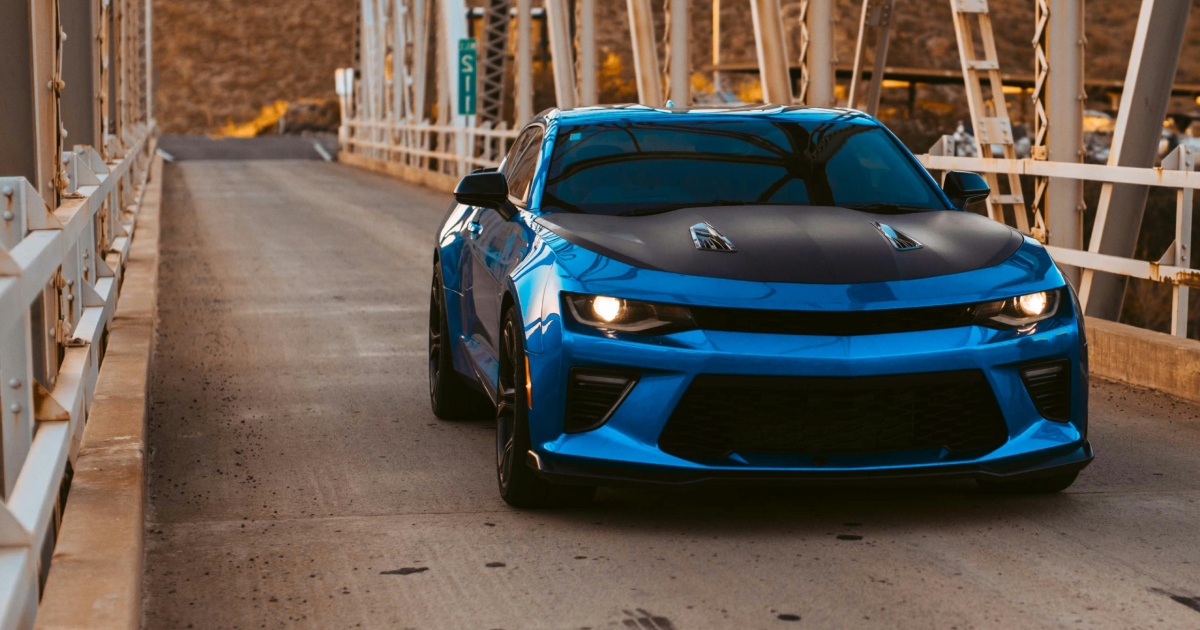 Blue sports car driving across a bridge with a black hood, daytime.