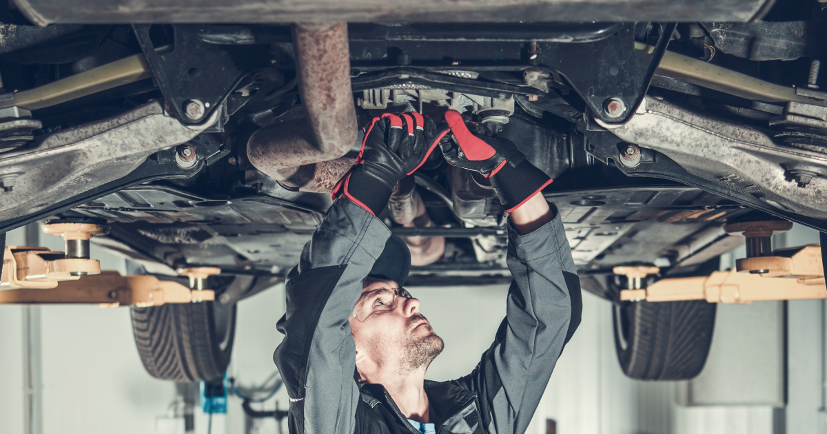 Mechanic works on a car's underside, wearing gloves, in a garage with the car lifted.