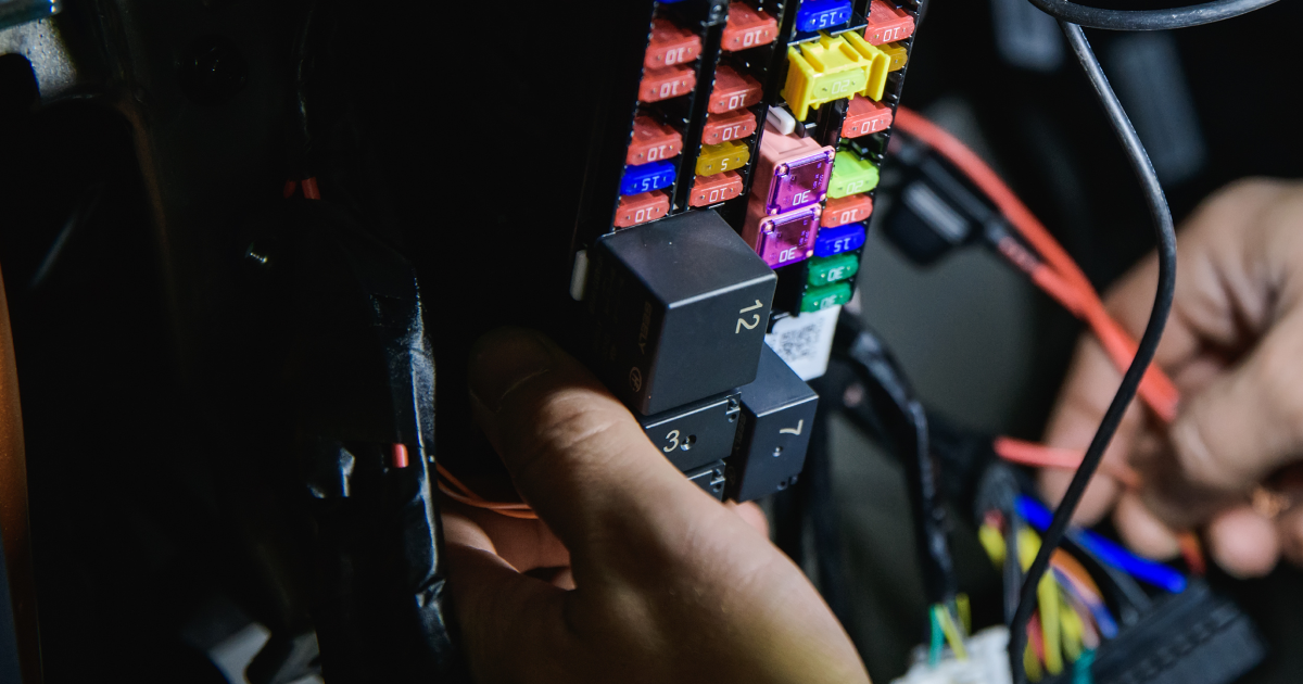 Person's hands near a car's fuse box, replacing a relay. Dark interior with colored fuses and wiring.