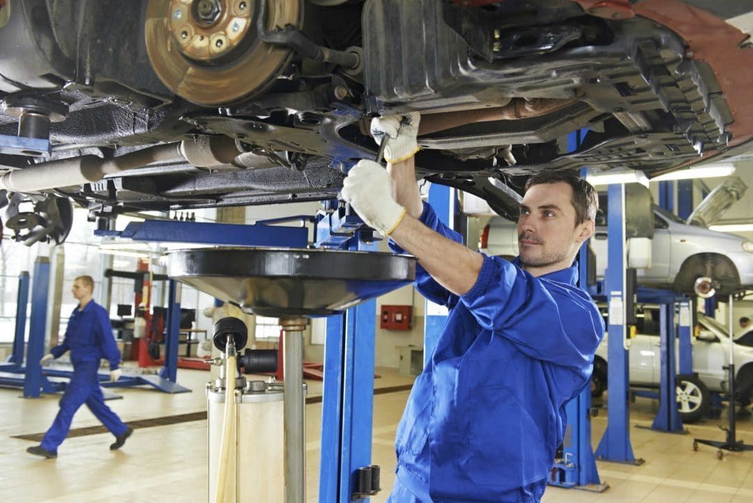 Mechanic in blue jumpsuit working under a car raised on a lift in a garage.