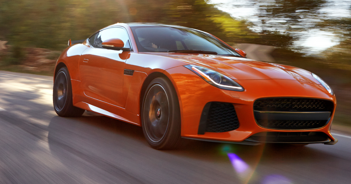 Orange Jaguar F-Type sports car speeding down a road, sunlight streaming through trees.