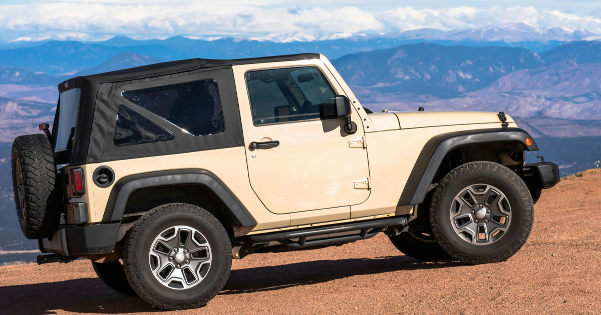 Beige Jeep Wrangler on a dirt road overlooking a mountain range with a black soft top.