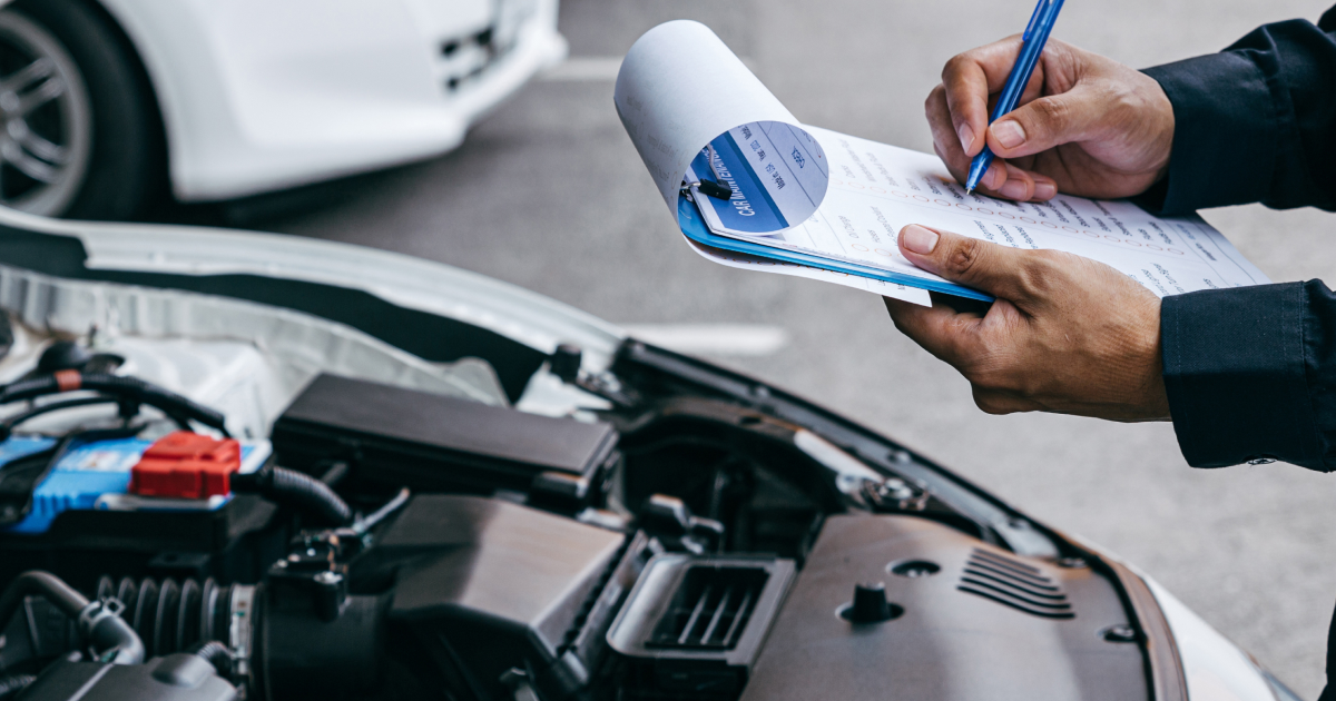 Mechanic inspecting car engine, writing on clipboard. White car in background, outdoors.
