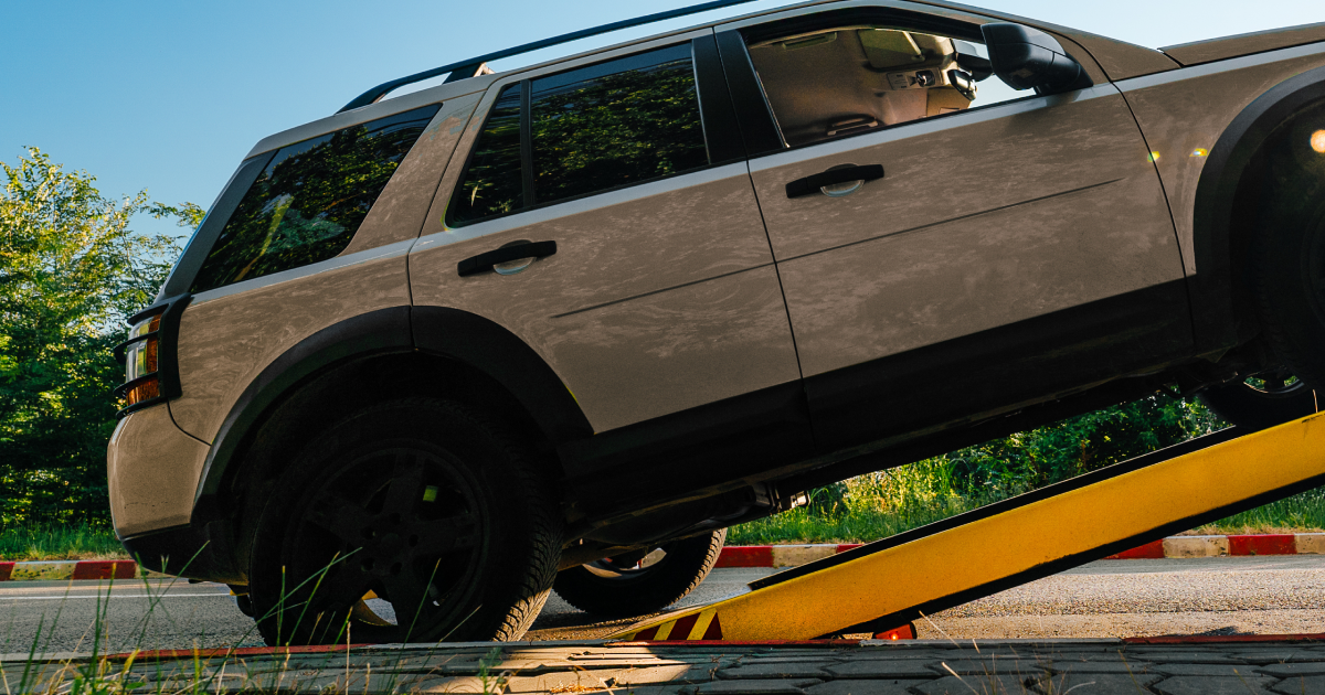 Beige SUV on a tow truck, angled upward. Black tires, blue sky, trees in the background.