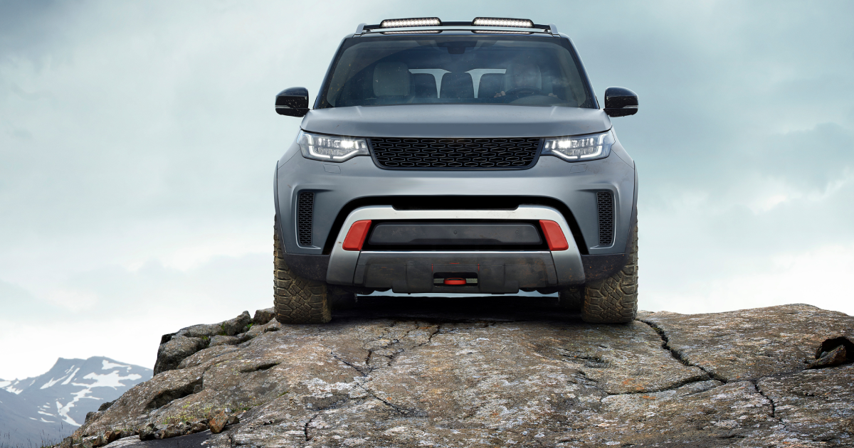 Gray Land Rover SUV on a rocky outcrop with a mountain range in the background. Red accents on the front.