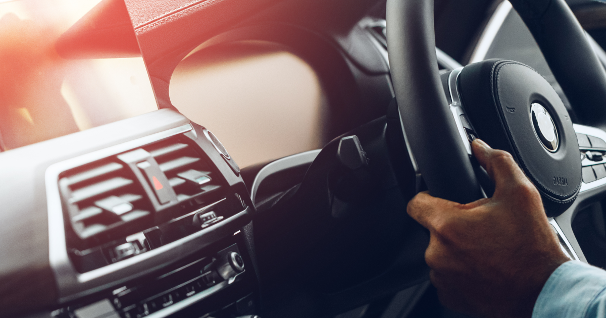 Person's hand on a car's steering wheel, driving. Interior view with dashboard and sunlight.