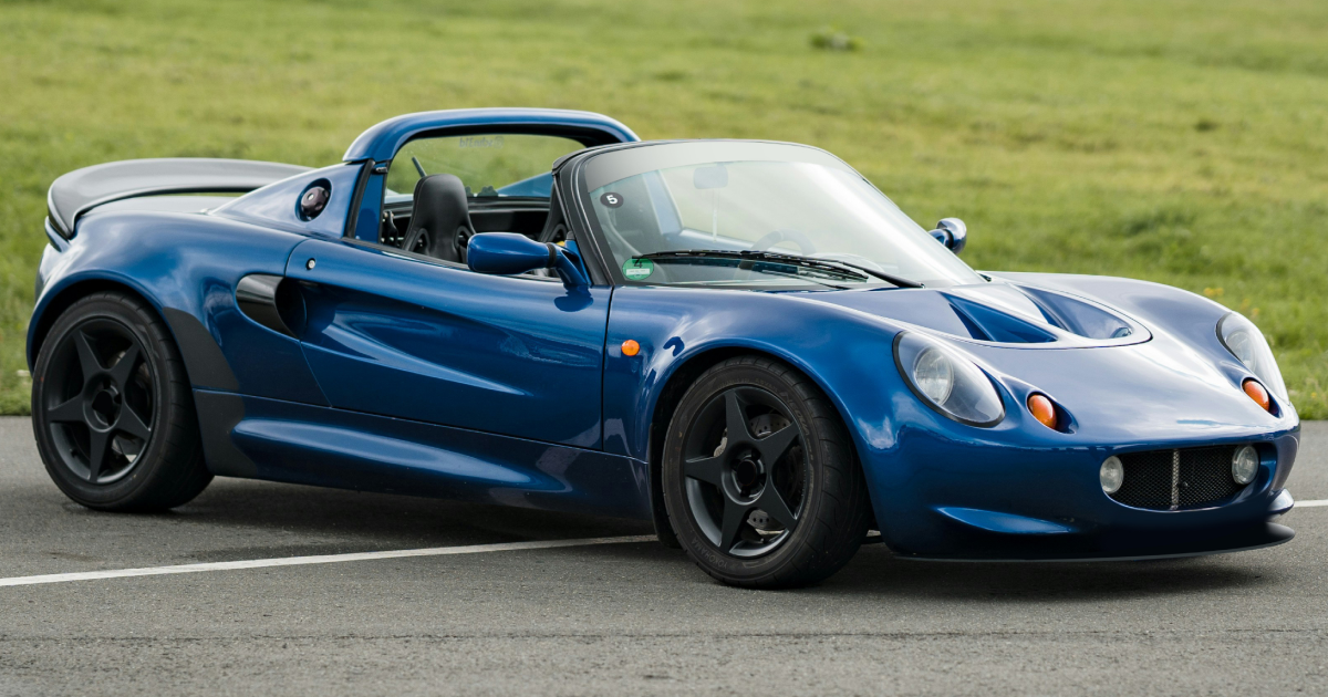 Blue Lotus Elise sports car on a track, black wheels, parked on asphalt with grass in the background.