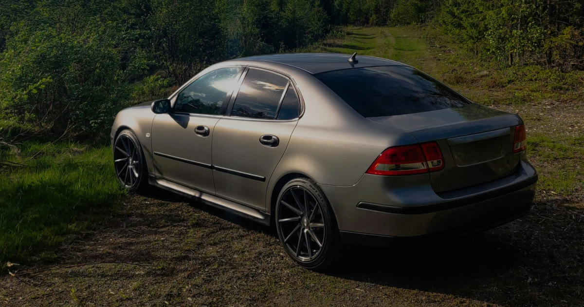 Tan Saab 9-3 sedan parked on a dirt road near greenery, showcasing its side profile with dark-colored wheels.