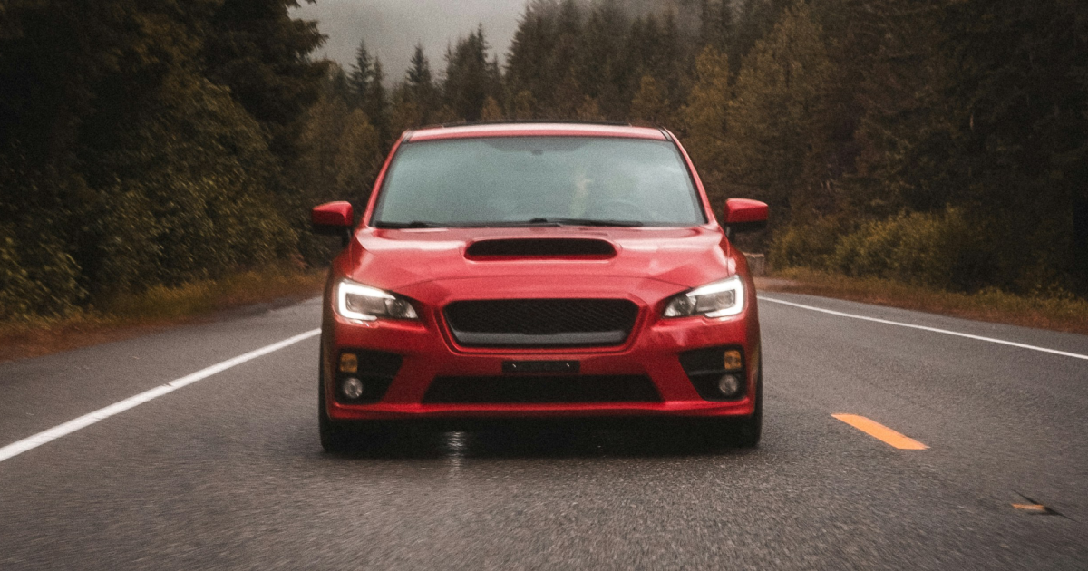Red Subaru WRX driving on a wet road through a forest, overcast weather.