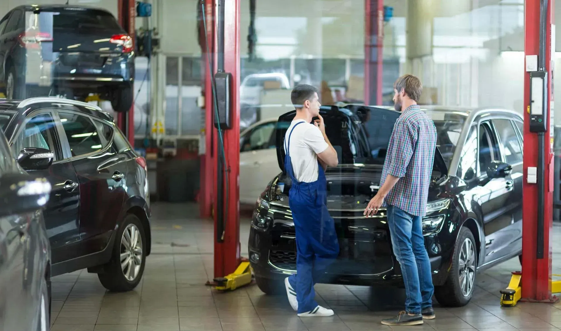 A mechanic and a customer discussing a car with its hood open inside a car repair shop.
