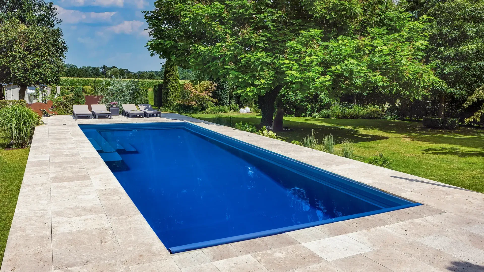Rectangular blue swimming pool surrounded by stone patio, green grass, and trees.