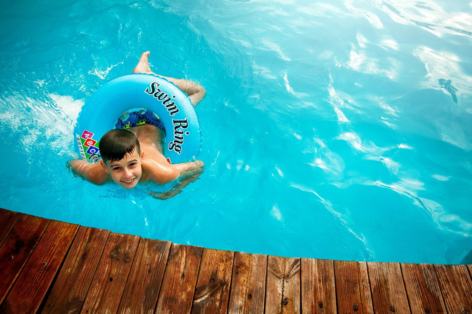 Boy in blue inner tube smiles in a bright blue pool; wooden deck in foreground.