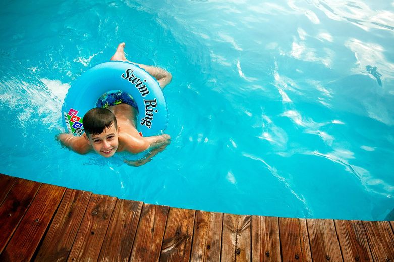 Boy in blue inner tube smiles in a bright blue pool; wooden deck in foreground.