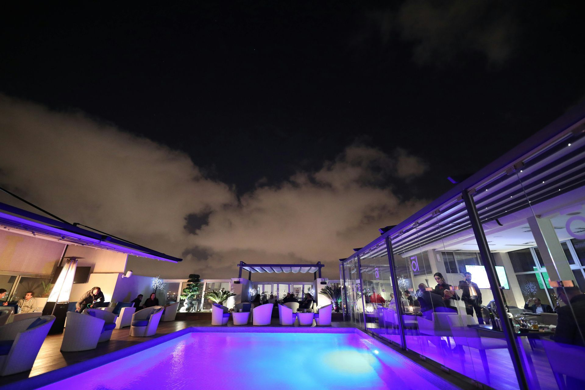 Rooftop pool and lounge at night. Lit in purple, people seated. Overcast sky.