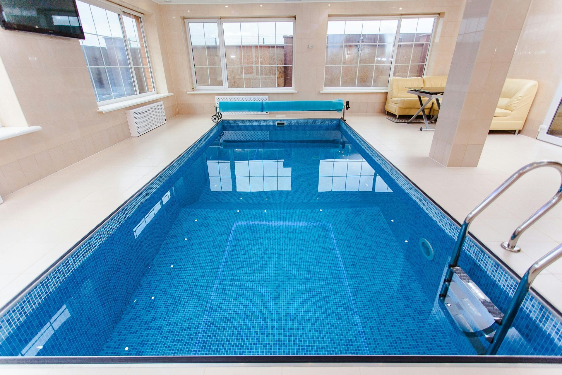 Indoor rectangular swimming pool with blue tiled surface, pool cover, and metal ladder. Windows line the wall.
