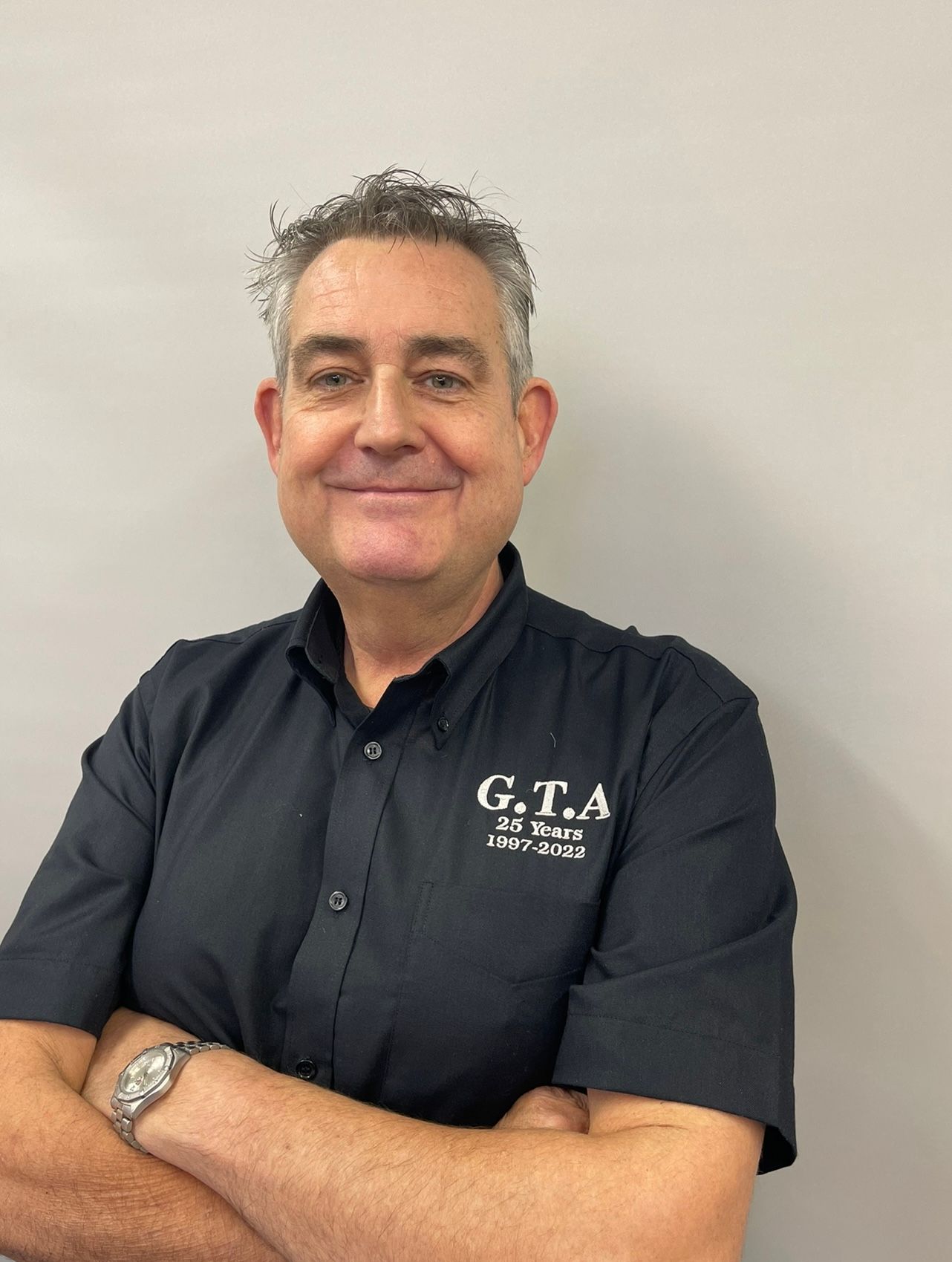 A man wearing a black shirt with the word g.t.a on it is standing with his arms crossed.