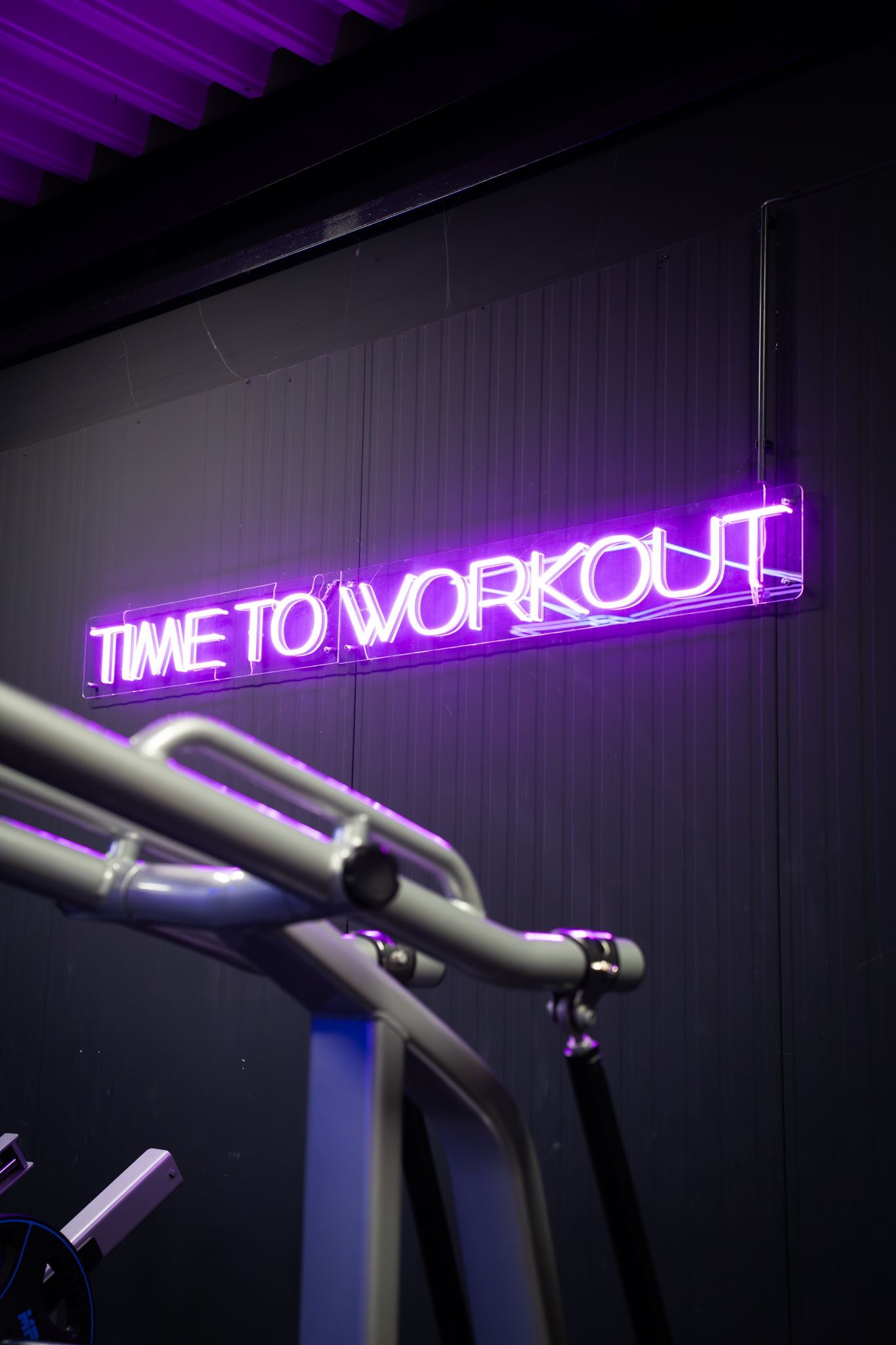 Neon purple sign reading “TIME TO WORKOUT” above gym equipment in a dark fitness room