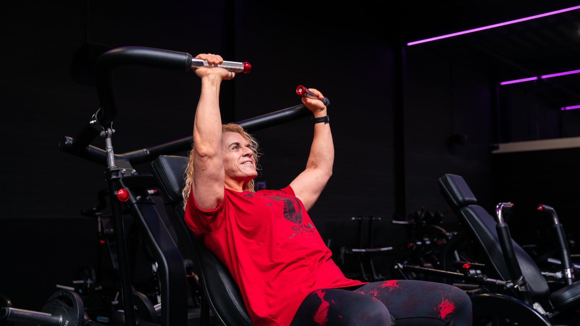 A person in red shirt doing a lat pulldown on gym equipment in a dark fitness center
